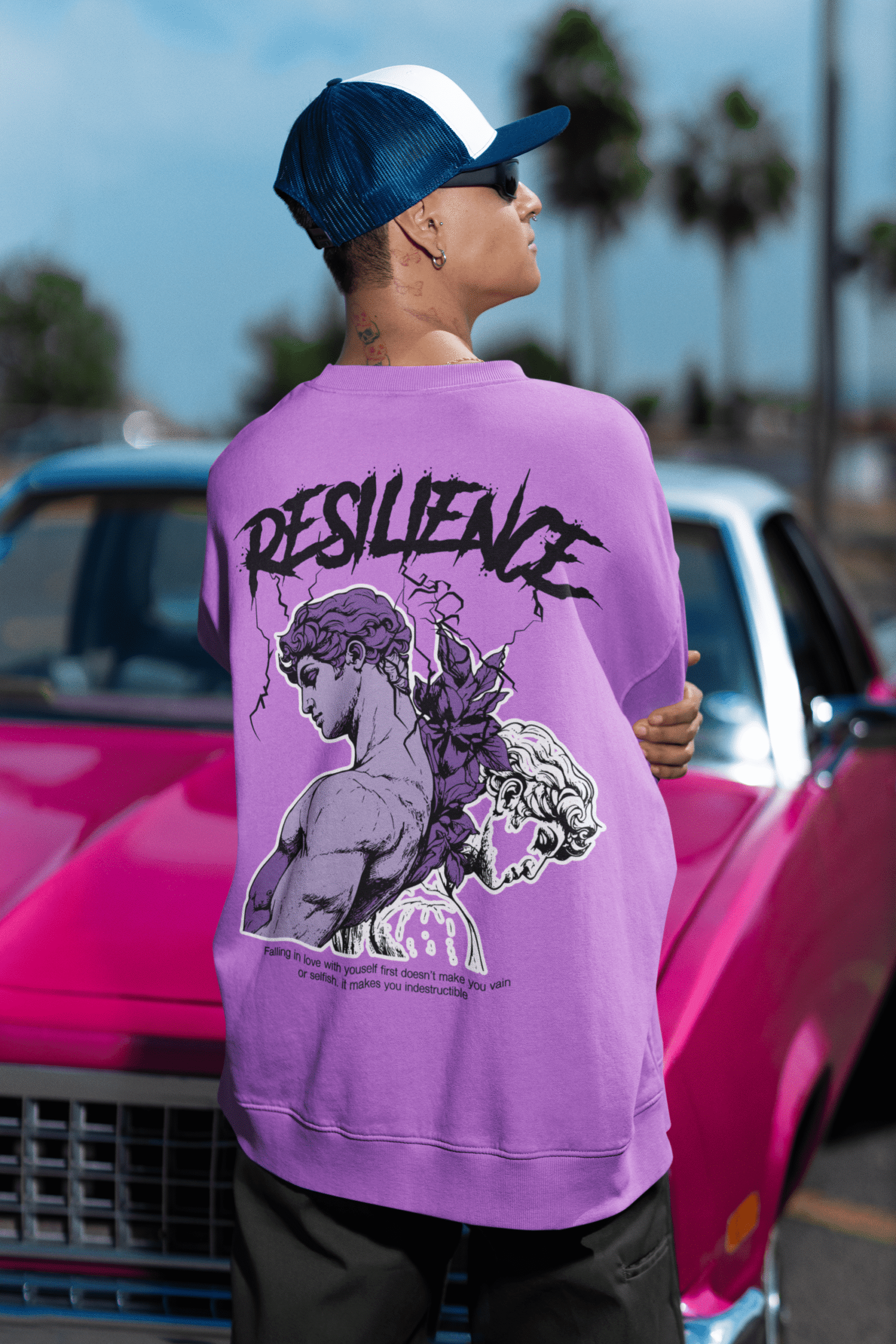 Model wearing a purple oversized sweatshirt with resilience graphic artwork on the back, styled with a vintage pink car outdoors.