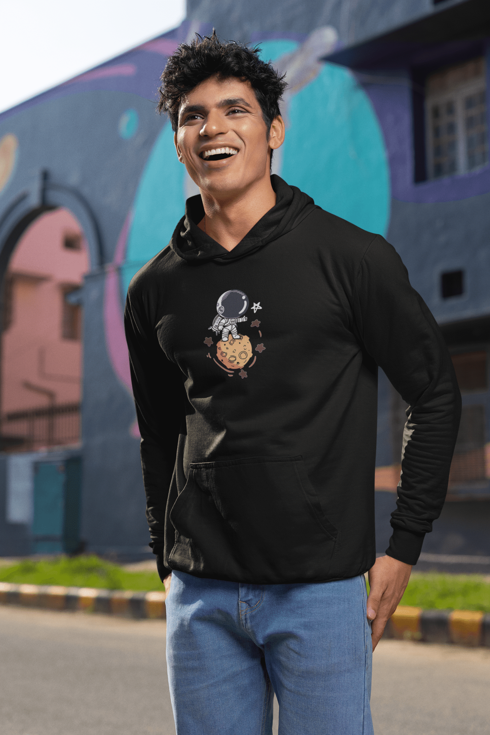 Man wearing a black astronaut graphic hoodie smiling outdoors near colorful street art