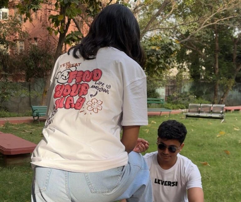 Two people wearing Leversi graphic T-shirts in an outdoor park setting; woman shows a “Find Your Way” back print while man wears a Leversi logo tee.