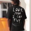 A man wearing a black T-shirt with large white handwritten text on the back that reads “LOOK MOM I CAN FLY,” along with a small graphic of a falling or flying figure near the bottom. He is outdoors, wearing light blue baggy jeans and a light-colored cap.