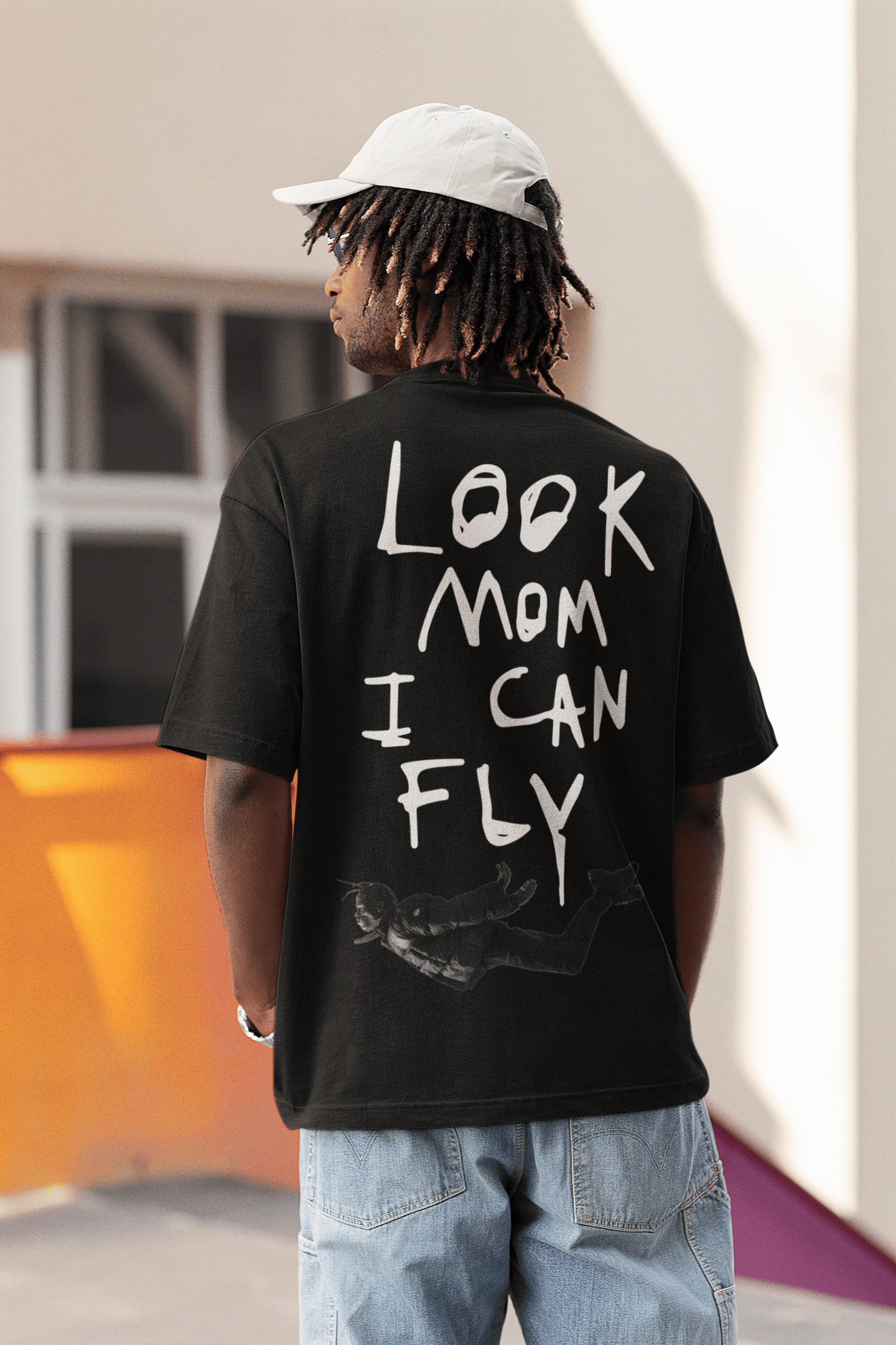 A man wearing a black T-shirt with large white handwritten text on the back that reads “LOOK MOM I CAN FLY,” along with a small graphic of a falling or flying figure near the bottom. He is outdoors, wearing light blue baggy jeans and a light-colored cap.