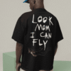 Man wearing a black oversized T-shirt with large handwritten white text on the back reading “LOOK MOM I CAN FLY,” along with a small flying figure graphic near the bottom. He is standing indoors on pastel green blocks, wearing a bright blue cap and beige shorts.