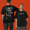Two models wearing black oversized Travis Scott–inspired graphic T-shirts; one showing a bold handwritten “LOOK MOM I CAN FLY” print with a flying figure on the back, and the other wearing a front graphic that reads “TRAVI$ SCOTT,” both posed against a solid orange background.