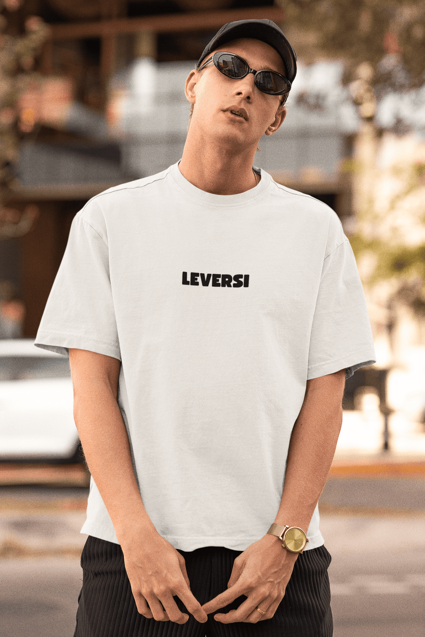 Man wearing a white Leversi logo T-shirt, sunglasses, and a cap while posing outdoors.