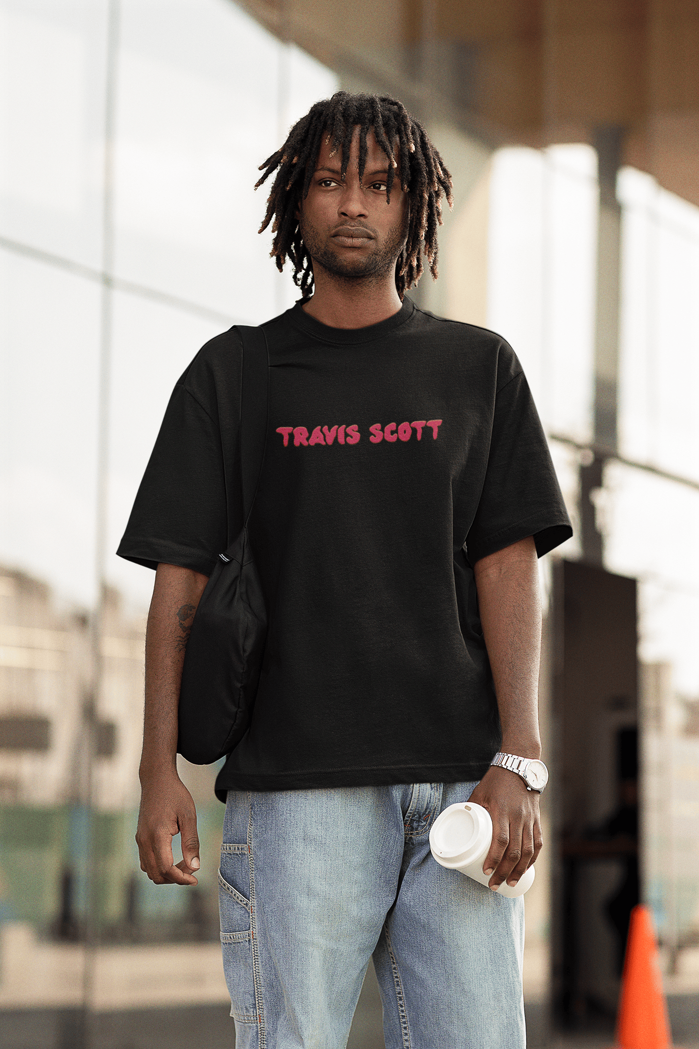 Man wearing a black Travis Scott T-shirt, light-wash jeans, and carrying a coffee cup in an outdoor urban setting.
