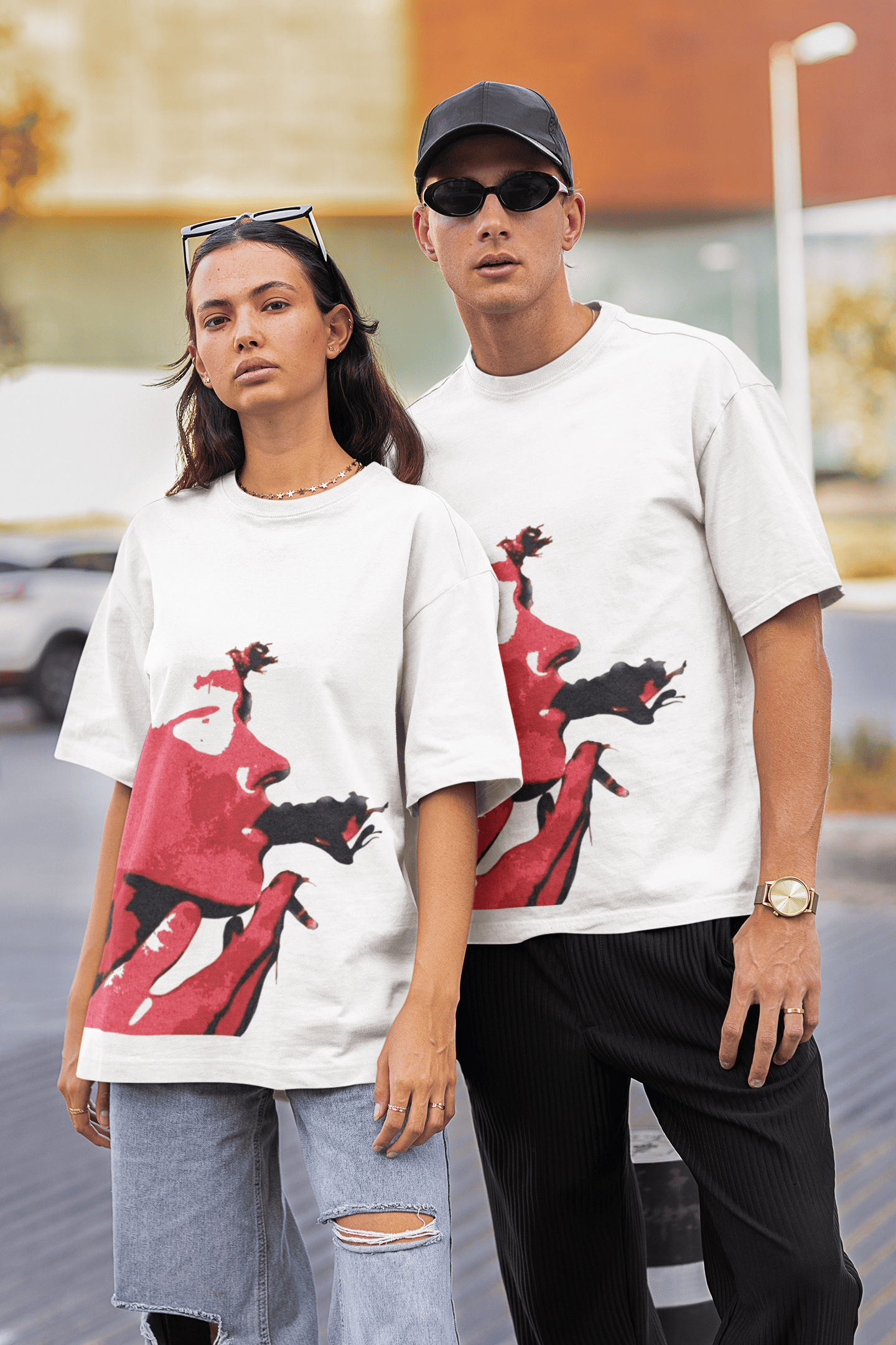 Man and woman wearing matching white graphic T-shirts with bold red face artwork in an outdoor urban setting.