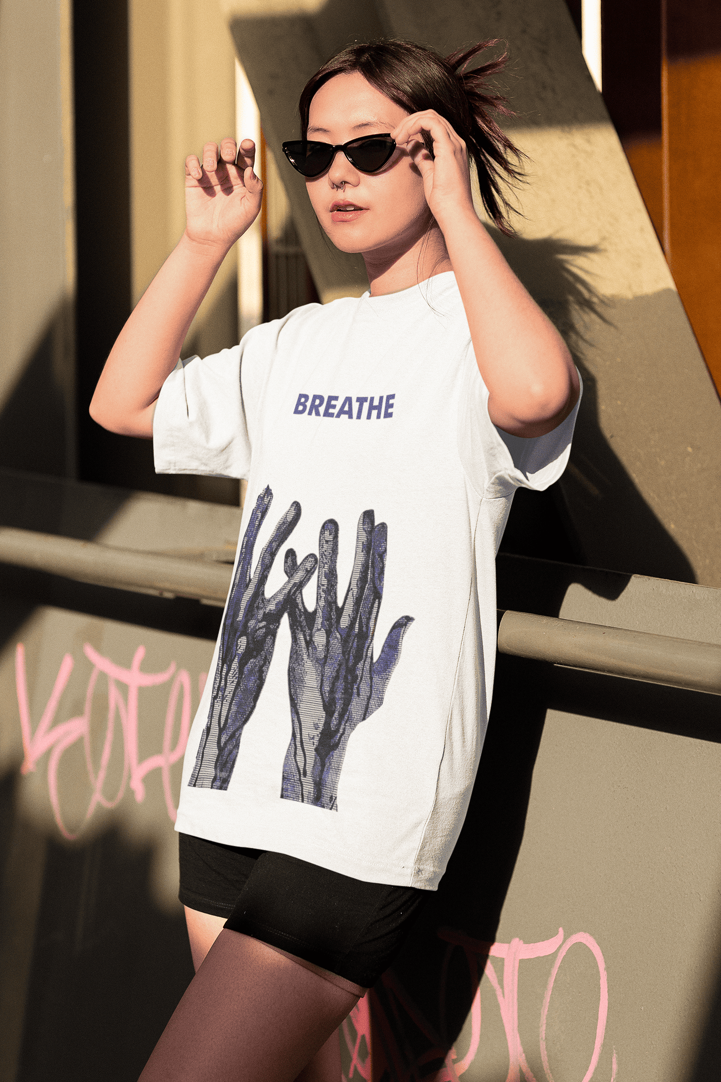 Woman wearing a white “BREATHE” T-shirt with blue abstract hand artwork, adjusting black cat-eye sunglasses while standing outdoors near a graffiti-marked structure.