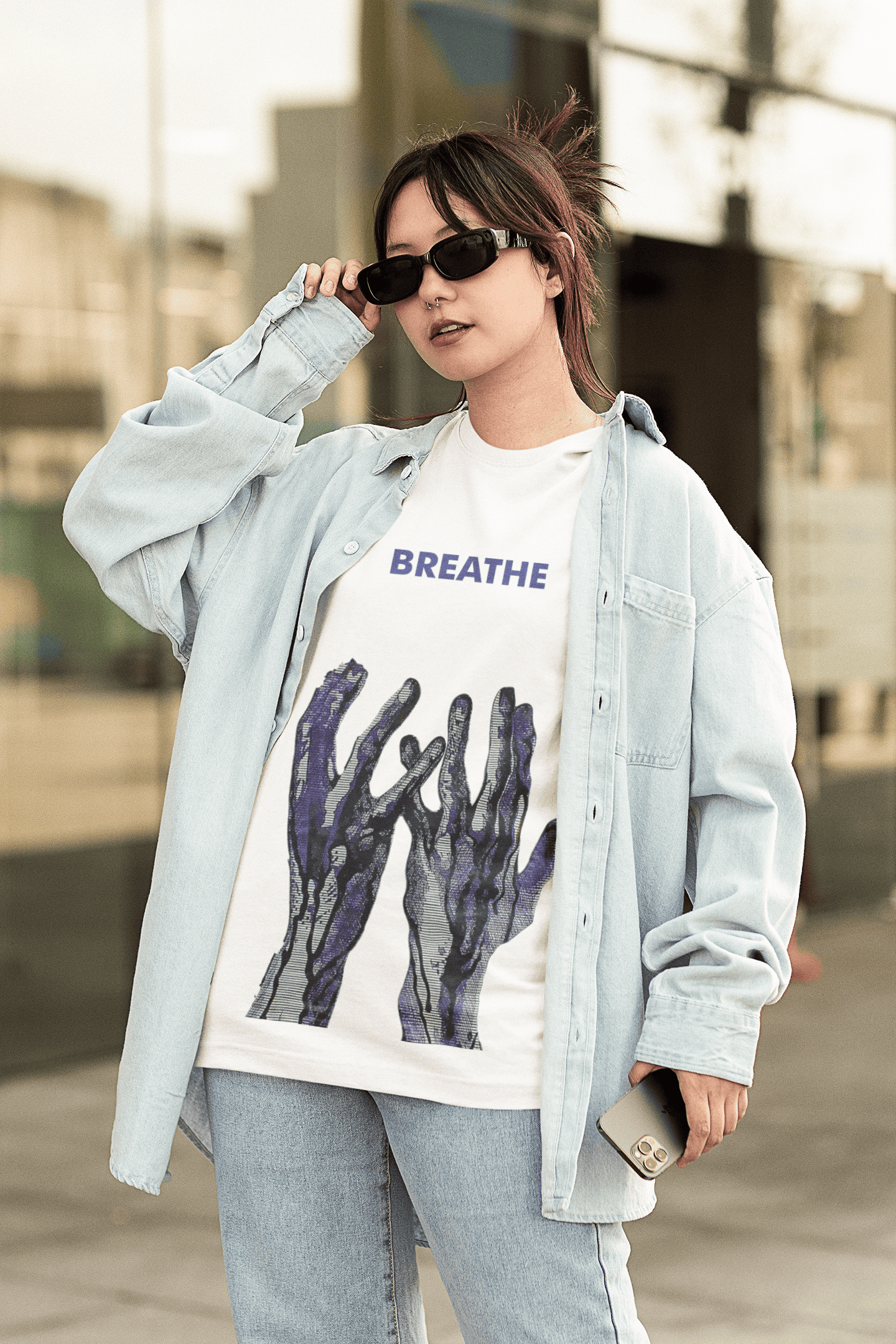 Woman wearing a white “BREATHE” graphic T-shirt with blue abstract hand design, styled with an oversized light denim shirt, jeans, and black sunglasses outdoors.