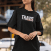 Woman wearing a black oversized T-shirt with a bold “TRAVIS SCOTT” graphic in white and red distortion effect, styled with sunglasses and light denim jeans outdoors.