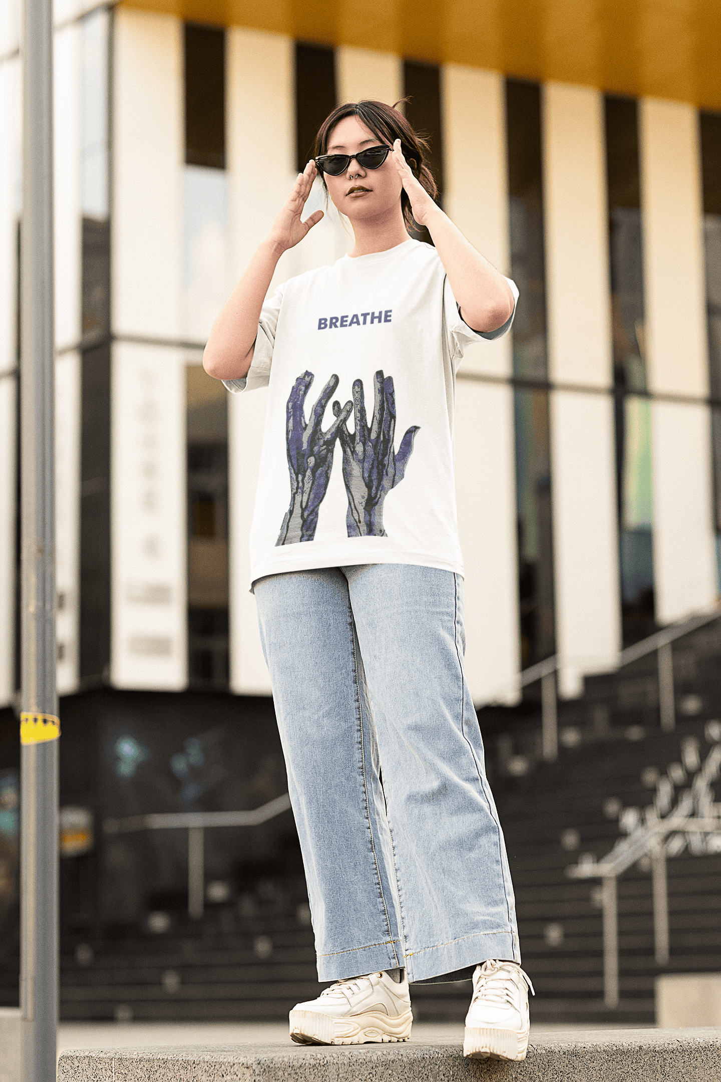 Woman wearing a white “BREATHE” graphic T-shirt with blue abstract hand design, paired with wide-leg denim jeans and sunglasses outdoors.