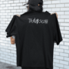 Woman holding up a black oversized T-shirt showing a white graffiti-style “TRAVI$ SCOTT” graphic on the back, standing outdoors against a white brick wall while wearing a black cap and sunglasses.