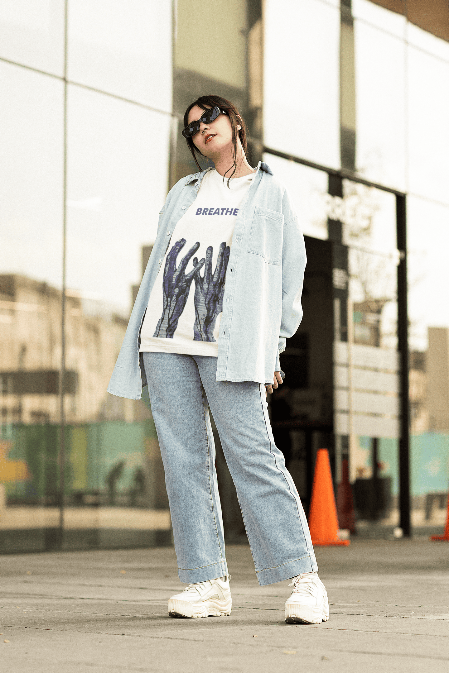 Woman wearing a white “BREATHE” graphic T-shirt with blue abstract hand design, layered with a denim shirt and jeans outdoors.